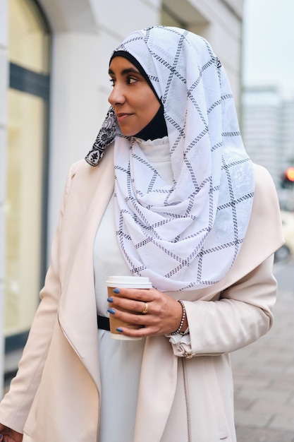 Junge attraktive arabische Frau im Hijab, die mit Kaffee durch die Stadtstraße geht