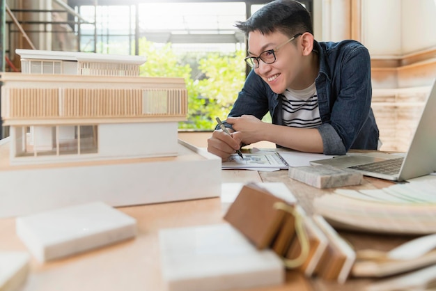 Junge attraktive asiatische Brille männlicher Architekt lächelt zuversichtlich, stolz darauf, ein neues Projekthausmodellglück asiatisch zu präsentieren, das mit Massenmodellstudie des Hausdesignprojekts arbeitet