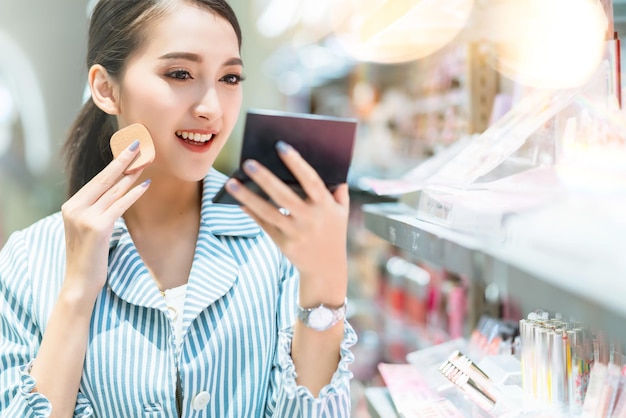 Junge attraktive asiatische Frau, die fröhlich ist, genießt es, Kosmetik im Einkaufszentrum zu wählen, unscharfer Bokeh-Hintergrund