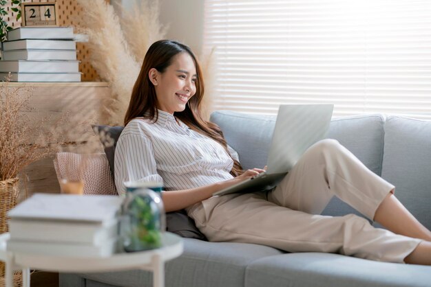 Junge attraktive asiatische frau, die zu hause arbeitet, benutzt laptop, der suchplan arbeitsprozessplanungsbericht mit freizeit überprüft, entspannen sie sich auf sofa im wohnzimmer mit morgenlicht friedlichem moment