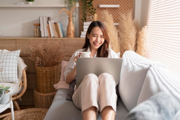 Junge attraktive asiatische frau, die zu hause arbeitet, benutzt laptop, der suchplan arbeitsprozessplanungsbericht mit freizeit überprüft, entspannen sie sich auf sofa im wohnzimmer mit morgenlicht friedlichem moment