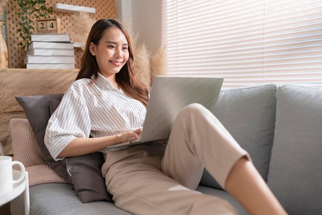 Junge attraktive asiatische frau, die zu hause arbeitet, benutzt laptop, der suchplan arbeitsprozessplanungsbericht mit freizeit überprüft, entspannen sie sich auf sofa im wohnzimmer mit morgenlicht friedlichem moment