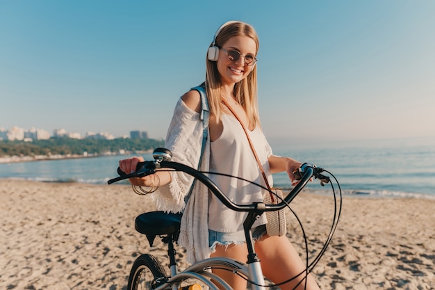 Junge attraktive blonde lächelnde Frau, die am Strand mit dem Fahrrad in den Kopfhörern geht, die Musik in der positiven glücklichen Stimmung hören