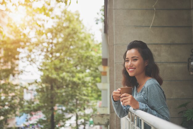 Junge attraktive Dame, die zur Kamera steht am Balkon und mit einer Tasse Tee kühlt lächelt