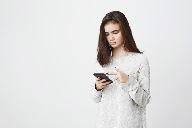 Junge attraktive europäische Frau, die Musik hört und Nachrichtenfeed in ihrem Smartphone mit konzentriertem Ausdruck scrollt. Frau sieht Live-Stream durch eine App
