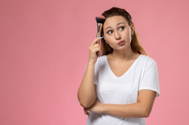 Junge attraktive Frau der Vorderansicht im weißen T-Shirt, das Make-up-Pinsel auf dem rosa Hintergrund hält