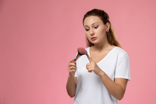 Junge attraktive Frau der Vorderansicht im weißen T-Shirt, das Make-up-Pinsel auf dem rosa Hintergrund hält