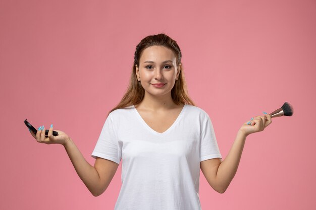 Junge attraktive Frau der Vorderansicht im weißen T-Shirt, das Make-up-Pinsel hält, der in die Kamera auf dem rosa Hintergrund schaut