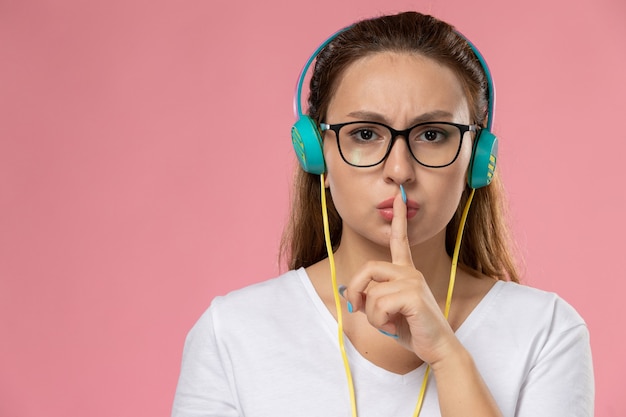 Kostenloses Foto junge attraktive frau der vorderansicht im weißen t-shirt, das musik hört, die stillezeichen auf dem rosa schreibtisch zeigt