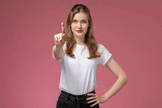 Junge attraktive Frau der Vorderansicht im weißen T-Shirt und in der schwarzen Hose, die mit Lächeln auf dem Farbfoto der weiblichen Wand des rosa Wandmodells posieren