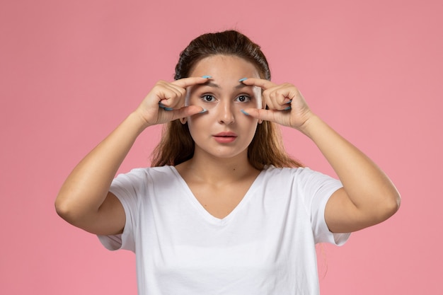 Kostenloses Foto junge attraktive frau der vorderansicht in der weißen t-shirt-aufstellung, die ihre augen auf dem rosa hintergrund zeigt