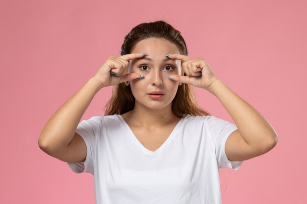 Junge attraktive Frau der Vorderansicht in der weißen T-Shirt-Aufstellung, die ihre Augen auf dem rosa Hintergrund zeigt