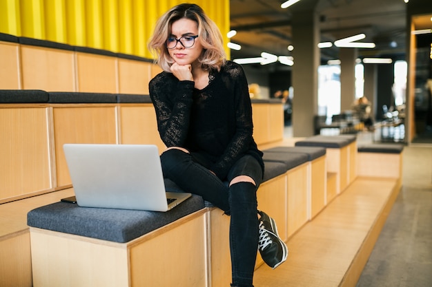 Junge attraktive Frau, die im Hörsaal sitzt, der an Laptop trägt Brille, modernes Auditorium, Studentenausbildung online, besorgtes Denken über Problem