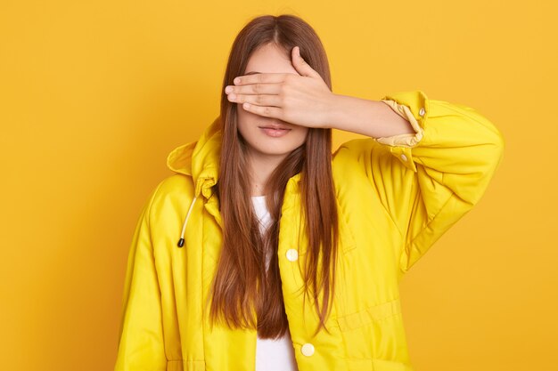 Junge attraktive Frau, die Jacke trägt, die ihre Augen mit Händen bedeckt, Frau mit langen Haaren, die gegen gelbe Wand steht, Dame versteckt sich vor ihrem Freund.