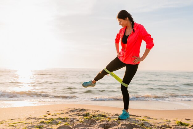 Junge attraktive Frau, die Sportübungen im Morgensonnenaufgang am Meeresstrand, gesunden Lebensstil tut, Musik auf Kopfhörern hörend, rosa Windbreakerjacke tragend macht, das Dehnen im Gummiband macht