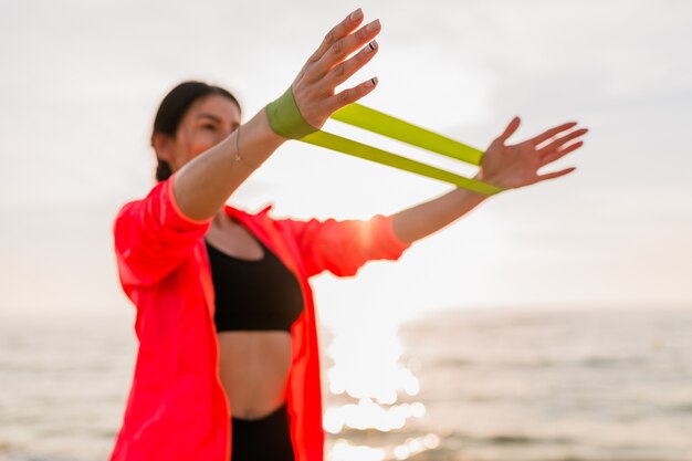 Junge attraktive Frau, die Sportübungen im Morgensonnenaufgang am Meeresstrand, gesunden Lebensstil tut, Musik auf Kopfhörern hörend, rosa Windbreakerjacke tragend macht, das Dehnen im Gummiband macht