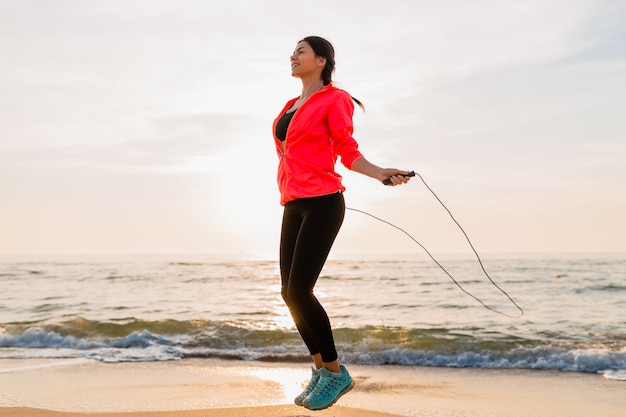 Junge attraktive Frau, die Sportübungen im Morgensonnenaufgang am Meeresstrand in der Sportkleidung, im gesunden Lebensstil, im Musikhören auf Kopfhörern, im Tragen der rosa Windjackejacke, springend im Springseil tut