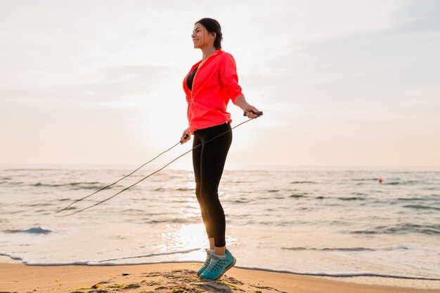 Junge attraktive Frau, die Sportübungen im Morgensonnenaufgang am Meeresstrand in der Sportkleidung, im gesunden Lebensstil, im Musikhören auf Kopfhörern, im Tragen der rosa Windjackejacke, springend im Springseil tut