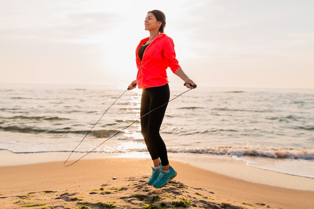 Junge attraktive Frau, die Sportübungen im Morgensonnenaufgang am Meeresstrand in der Sportkleidung, im gesunden Lebensstil, im Musikhören auf Kopfhörern, im Tragen der rosa Windjackejacke, springend im Springseil tut