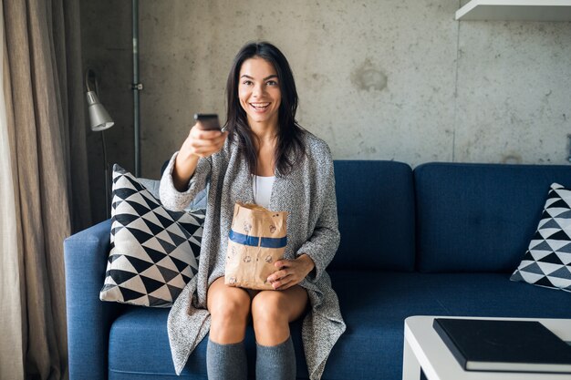 Junge attraktive Frau im lässigen Outfit, das zu Hause Filme im Fernsehen hält, Fernbedienung hält, Kanäle wechselt, Popcorn isst, Freizeit, Spaß hat, lächelnd auf Sofa sitzend, fröhlich, glücklich