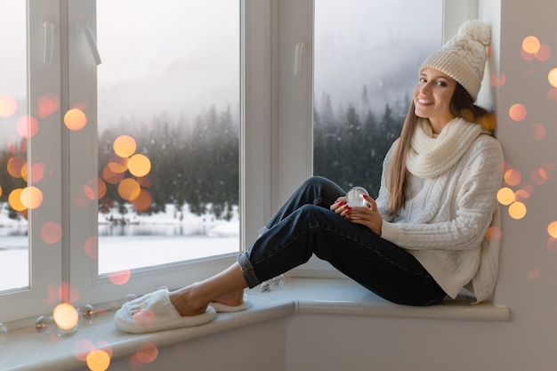 Junge attraktive Frau im stilvollen weißen Strickpullover, im Schal und in der Mütze, die zu Hause auf der Fensterbank zu Weihnachten sitzen und Glasschneekugelgeschenkdekoration, Winterwaldansicht, Lichter bokeh halten