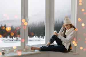 Kostenloses Foto junge attraktive frau im stilvollen weißen strickpullover, im schal und in der mütze, die zu hause auf der fensterbank zu weihnachten sitzen und glasschneekugelgeschenkdekoration, winterwaldansicht, lichter bokeh halten