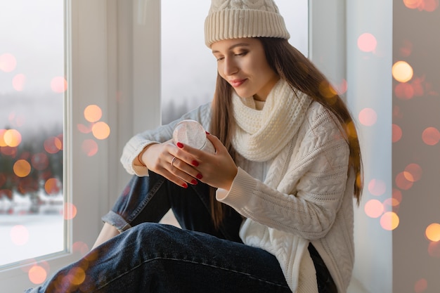 Junge attraktive Frau im stilvollen weißen Strickpullover, im Schal und in der Mütze, die zu Hause auf der Fensterbank zu Weihnachten sitzen und Glasschneekugelgeschenkdekoration, Winterwaldansicht, Lichter bokeh halten