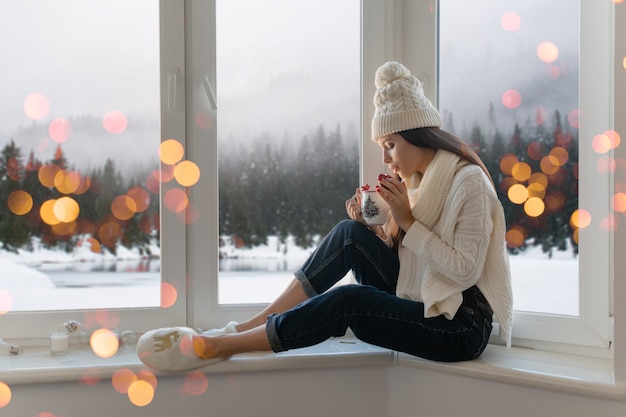 Junge attraktive Frau im stilvollen weißen Strickpullover, im Schal und in der Mütze, die zu Hause auf Fensterbank an Weihnachten halten Tasse halten, die heißen Tee trinkt, Winterwaldhintergrundansicht, Lichter bokeh