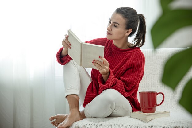 Junge attraktive Frau in einem gemütlichen roten Pullover liest ein Buch, während sie zu Hause mit einer Tasse Getränk auf dem Sofa sitzt.