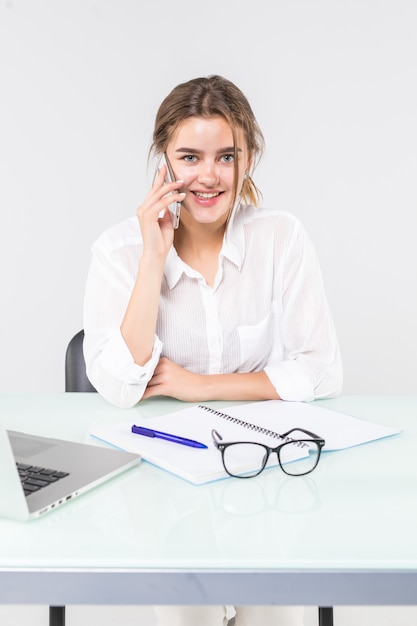 Junge attraktive Frau in Pastell-Freizeitkleidung, die auf Handy spricht, sitzen Arbeit am Schreibtisch mit PC-Laptop lokalisiert auf grauem Hintergrund.