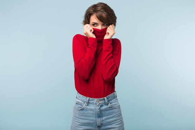 Junge attraktive Frau mit dunklen kurzen Haaren, die das Gesicht in einem roten Pullover versteckt, während sie nachdenklich über blauem Hintergrund zur Seite schaut