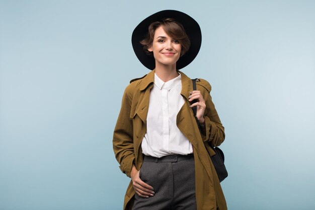 Junge attraktive Frau mit dunklen kurzen Haaren in Trenchcoat und Hut mit Rucksack, die glücklich in die Kamera schaut, isoliert auf blauem Hintergrund