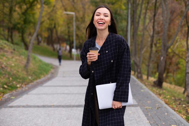 Junge attraktive fröhliche Frau im Mantel mit Kaffee zum Mitnehmen und Laptop, die fröhlich im Herbstpark spazieren geht