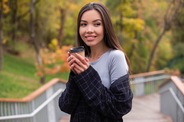 Junge attraktive glückliche Frau im Mantel mit Kaffee zum Mitnehmen, der verträumt im Stadtpark wegschaut