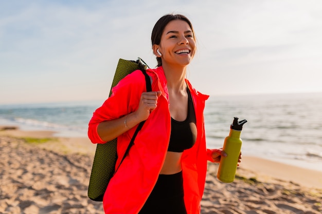 Junge attraktive lächelnde Frau, die Sport im Morgensonnenaufgang am Meeresstrand tut, der Yogamatte und Flasche Wasser hält, gesunder Lebensstil, Musik auf Kopfhörern hörend, rosa Windjacke tragend