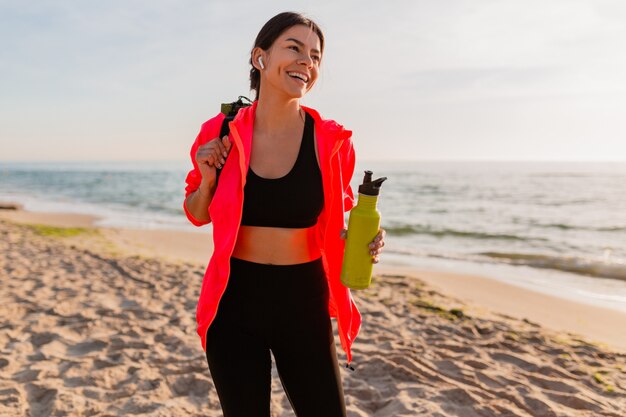 Junge attraktive lächelnde Frau, die Sport im Morgensonnenaufgang am Meeresstrand tut, der Yogamatte und Flasche Wasser hält, gesunder Lebensstil, Musik auf Kopfhörern hörend, rosa Windjacke tragend