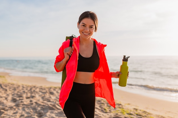 Junge attraktive lächelnde Frau, die Sport im Morgensonnenaufgang am Meeresstrand tut, der Yogamatte und Flasche Wasser hält, gesunder Lebensstil, Musik auf Kopfhörern hörend, rosa Windjacke tragend