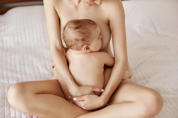 Junge attraktive nackte Mutter, die stillt und ihr neugeborenes Baby lächelnd sitzend auf Bett zu Hause umarmt.