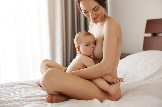 Junge attraktive nackte Mutter, die stillt und ihr neugeborenes Baby lächelnd sitzend auf Bett zu Hause umarmt.