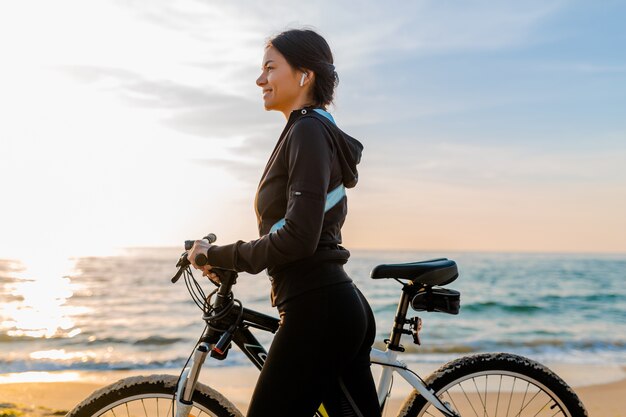 Junge attraktive schlanke Frau, die Fahrrad reitet, Sport am Morgensonnenaufgangstrand in Sportfitness-Sportkleidung, aktiven gesunden Lebensstil, lächelnd glücklich, Spaß zu haben