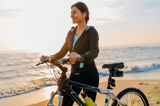 Junge attraktive schlanke Frau, die Fahrrad reitet, Sport am Morgensonnenaufgangstrand in Sportfitness-Sportkleidung, aktiven gesunden Lebensstil, lächelnd glücklich, Spaß zu haben