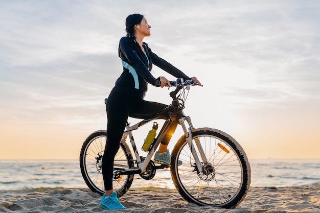 Junge attraktive schlanke Frau, die Fahrrad reitet, Sport im Morgensonnenaufgang-Sommerstrand in Sportfitnesskleidung, aktiver gesunder Lebensstil, lächelnd glücklich, Spaß zu haben