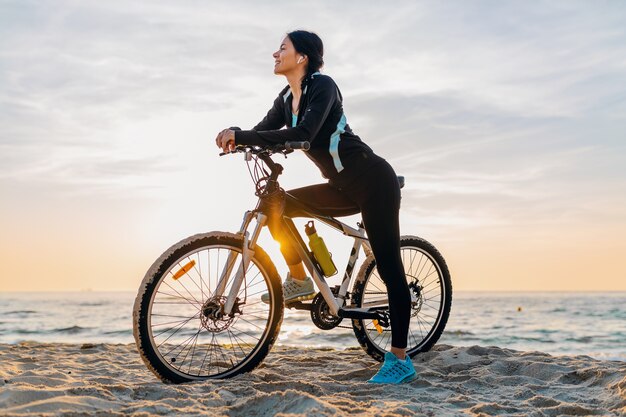 Junge attraktive schlanke Frau, die Fahrrad reitet, Sport im Morgensonnenaufgang-Sommerstrand in Sportfitnesskleidung, aktiver gesunder Lebensstil, lächelnd glücklich, Spaß zu haben