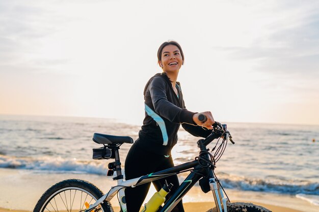 Junge attraktive schlanke Frau, die Fahrrad reitet, Sport im Morgensonnenaufgang-Sommerstrand in Sportfitnesskleidung, aktiver gesunder Lebensstil, lächelnd glücklich, Spaß zu haben