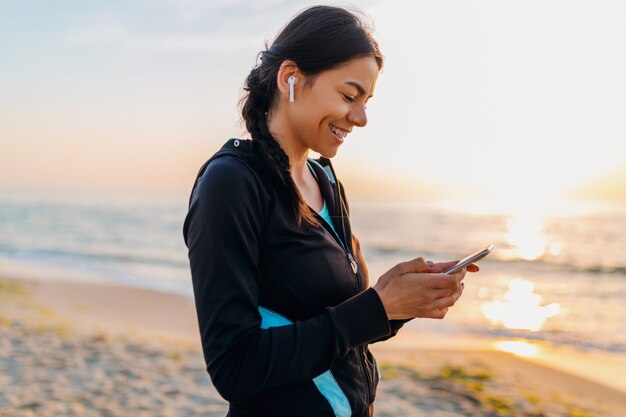 Junge attraktive schlanke Frau, die Sportübungen am Morgensonnenaufgangstrand in der Sportkleidung, im gesunden Lebensstil, im Musikhören auf drahtlosen Kopfhörern hält, die Smartphone halten, glücklich lächelnd Spaß haben