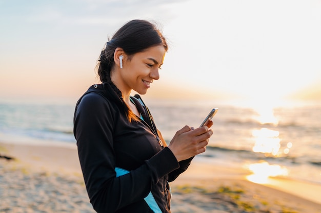 Junge attraktive schlanke Frau, die Sportübungen am Morgensonnenaufgangstrand in der Sportkleidung, im gesunden Lebensstil tut, Musik auf drahtlosen Kopfhörern haltend Smartphone hält und glücklich lächelt