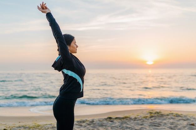 Junge attraktive schlanke Frau, die Sportübungen am Morgensonnenaufgangstrand in Sportkleidung, gesundem Lebensstil, Musik auf Kopfhörern hörend macht, das Strecken macht