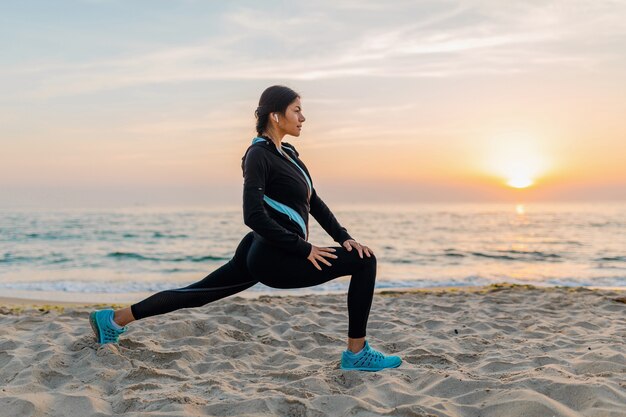 Junge attraktive schlanke Frau, die Sportübungen am Morgensonnenaufgangstrand in Sportkleidung, gesundem Lebensstil, Musik auf Kopfhörern hörend macht, das Strecken macht