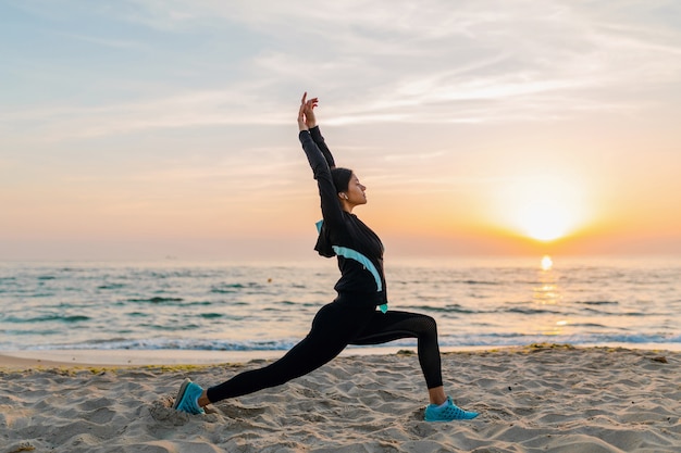 Junge attraktive schlanke Frau, die Sportübungen am Morgensonnenaufgangstrand in Sportkleidung, gesundem Lebensstil, Musik auf Kopfhörern hörend macht, das Strecken macht