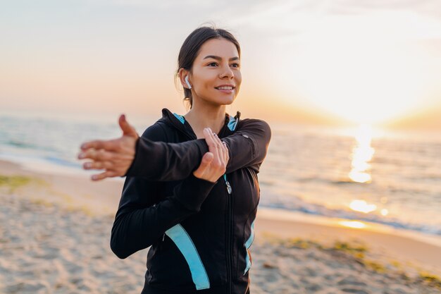 Junge attraktive schlanke Frau, die Sportübungen am Morgensonnenaufgangstrand in Sportkleidung, gesundem Lebensstil, Musik auf Kopfhörern hörend macht, das Strecken macht